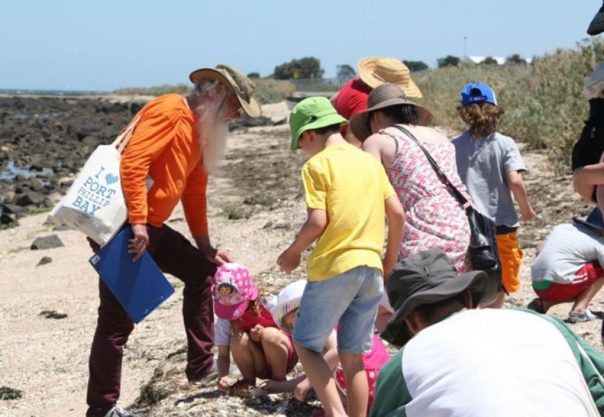 A group of adults and children on a beach. All wearing sunhats and brightly coloured clothes. All are focused on the sand, looking down. One adult is holding a blue clip board and a cream coloured bag that says "I heart Port Phillip Bay" in blue.