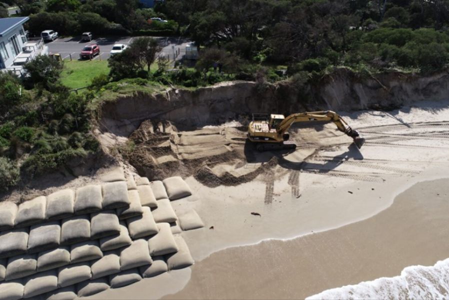 A construction site on the beach. There is a digger and piles of sand bags. 