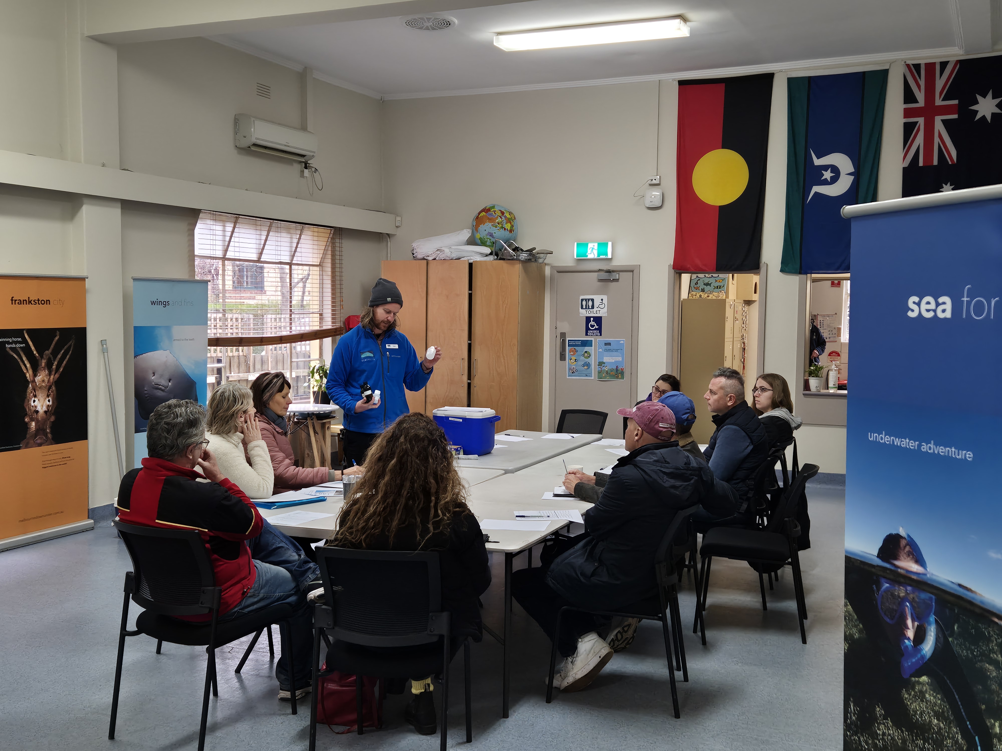 Group of people gathered around a table with 1 person standing and addressing the group