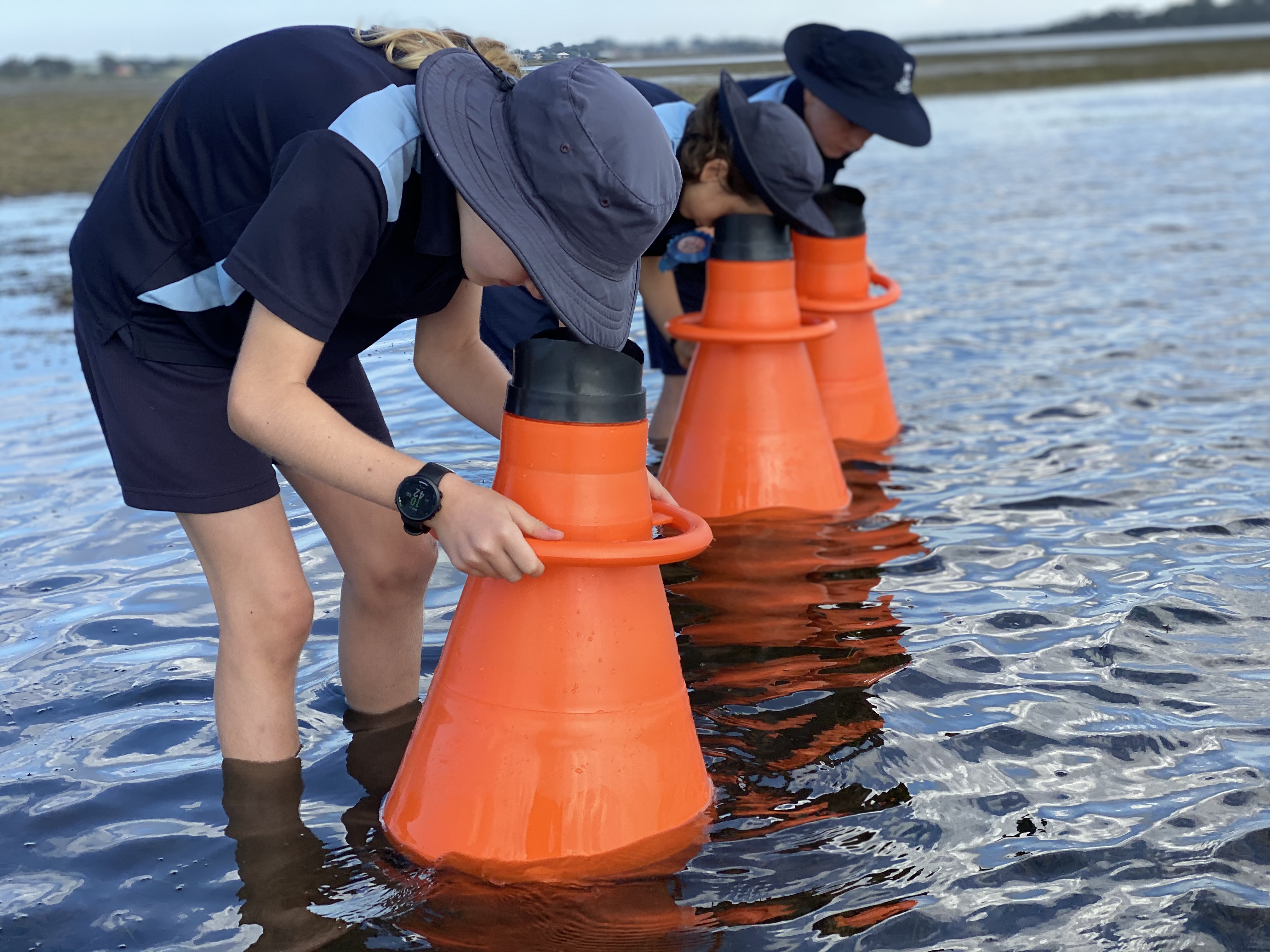 Image is a photograph of 3 people standing shin-deep in water each bending down to look through a piece of equipment in front of them into the water