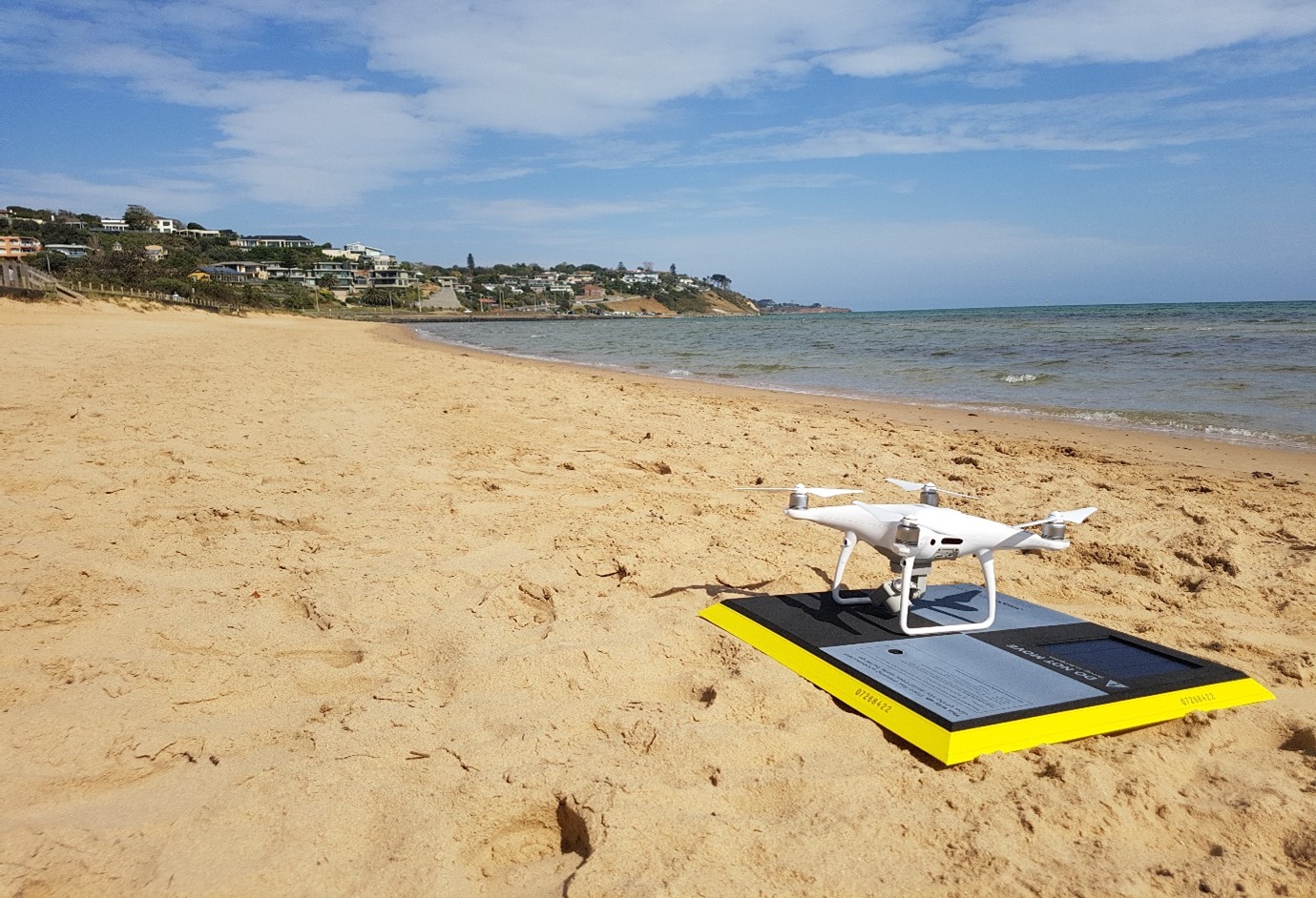 A drone is parked on the beach, nearby the ocean. Melbourne city is in the background.